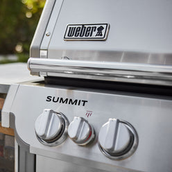 Close-up of a stainless steel gas grill’s lid handle and control panel, with two large burner knobs and a smaller infrared broiler knob marked by a red heat symbol.