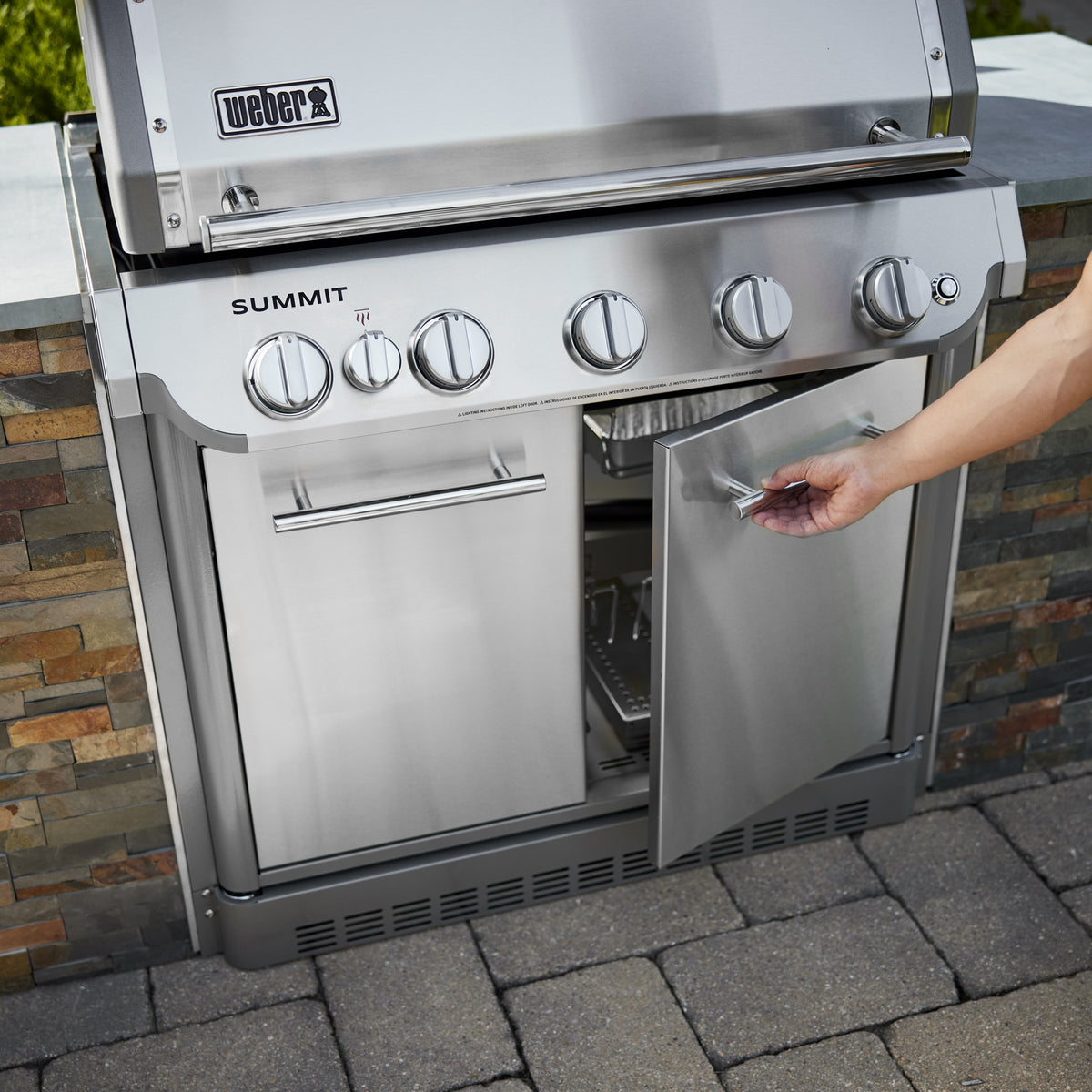 A hand opens the right cabinet door of a stainless steel built-in gas grill installed in a stone outdoor kitchen island.