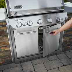 A hand opens the right cabinet door of a stainless steel built-in gas grill installed in a stone outdoor kitchen island.