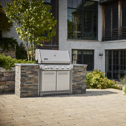 Stainless steel built-in gas grill with a closed lid and double-door cabinet installed in a stone outdoor kitchen island on a paved patio.