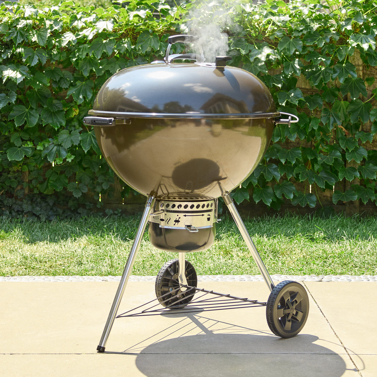 Smoke rising from the lid vent of the Weber 26” Master-Touch Charcoal Grill in Smoke Grey on a sunny patio, with an enclosed ash catcher, two wheels, and a wire storage shelf.