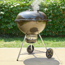Smoke rising from the lid vent of the Weber 26” Master-Touch Charcoal Grill in Smoke Grey on a sunny patio, with an enclosed ash catcher, two wheels, and a wire storage shelf.