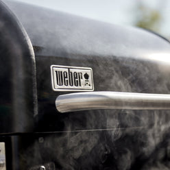 Close-up of the Weber logo and stainless steel lid handle on the pellet grill with smoke rising.