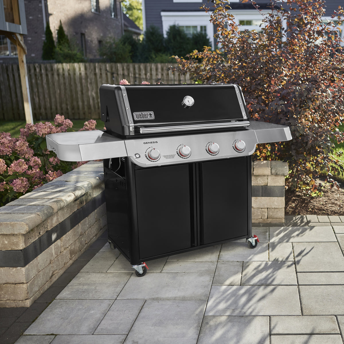On a stone patio, the Weber Genesis E-415 Gas Grill features two side tables, four burner controls, a closed black lid with a thermometer, and a double-door storage cabinet.