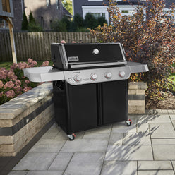 On a stone patio, the Weber Genesis E-415 Gas Grill features two side tables, four burner controls, a closed black lid with a thermometer, and a double-door storage cabinet.