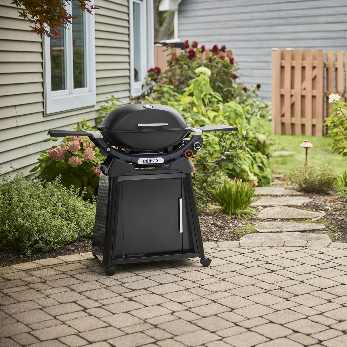 Midnight Black Gas Grill on a wheeled cabinet cart on a paved backyard patio, with a closed domed lid, built-in thermometer, and extended side tables.