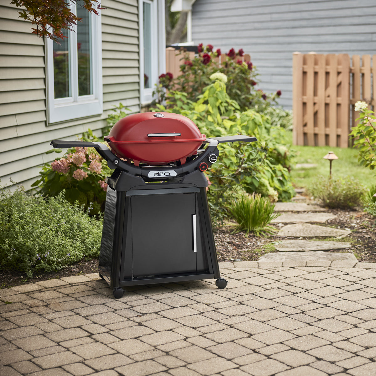 Flame Red Gas Grill on a wheeled cabinet cart on a paved backyard patio, with a closed domed lid, built-in thermometer, and extended side tables.