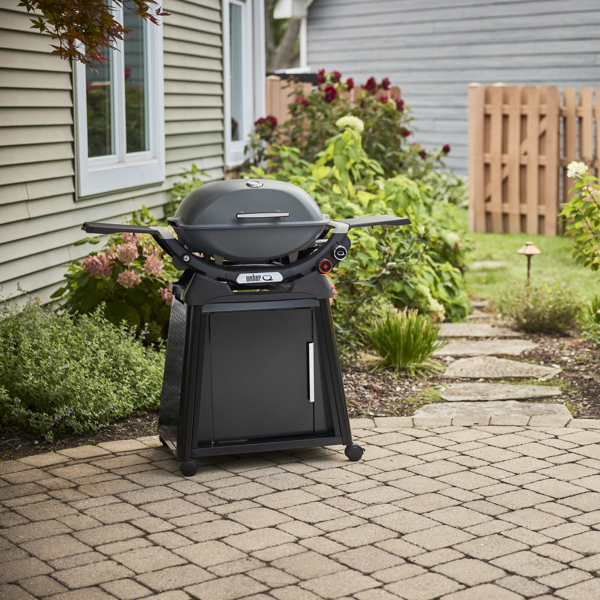 Charcoal Gray Gas Grill on a wheeled cabinet cart on a paved backyard patio, with a closed domed lid, built-in thermometer, and extended side tables.