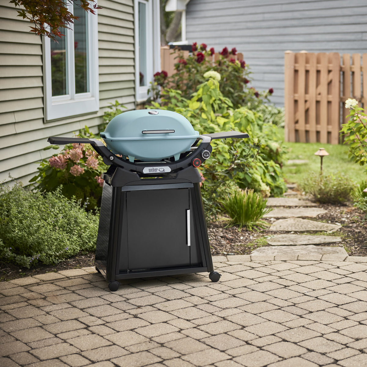 Sky Blue Gas Grill on a wheeled cabinet cart on a paved backyard patio, with a closed domed lid, built-in thermometer, and extended side tables.