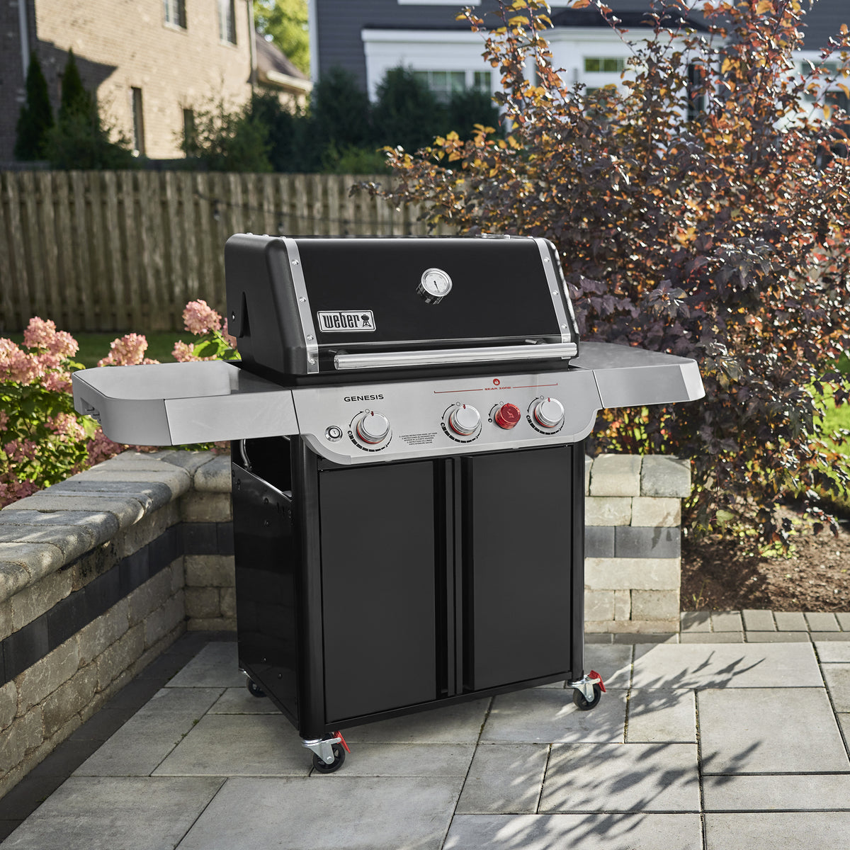 Weber Genesis E-325W Titanium Edition gas grill ready for outdoor cooking on a stone backyard patio surrounded by flowers and trees.