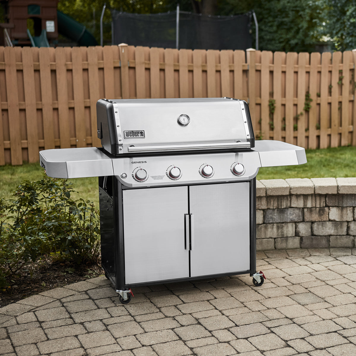 Stainless steel Weber Genesis S-415 Gas Grill on a backyard patio, showing the enclosed cabinet and locking caster wheels for easy movement and secure placement.