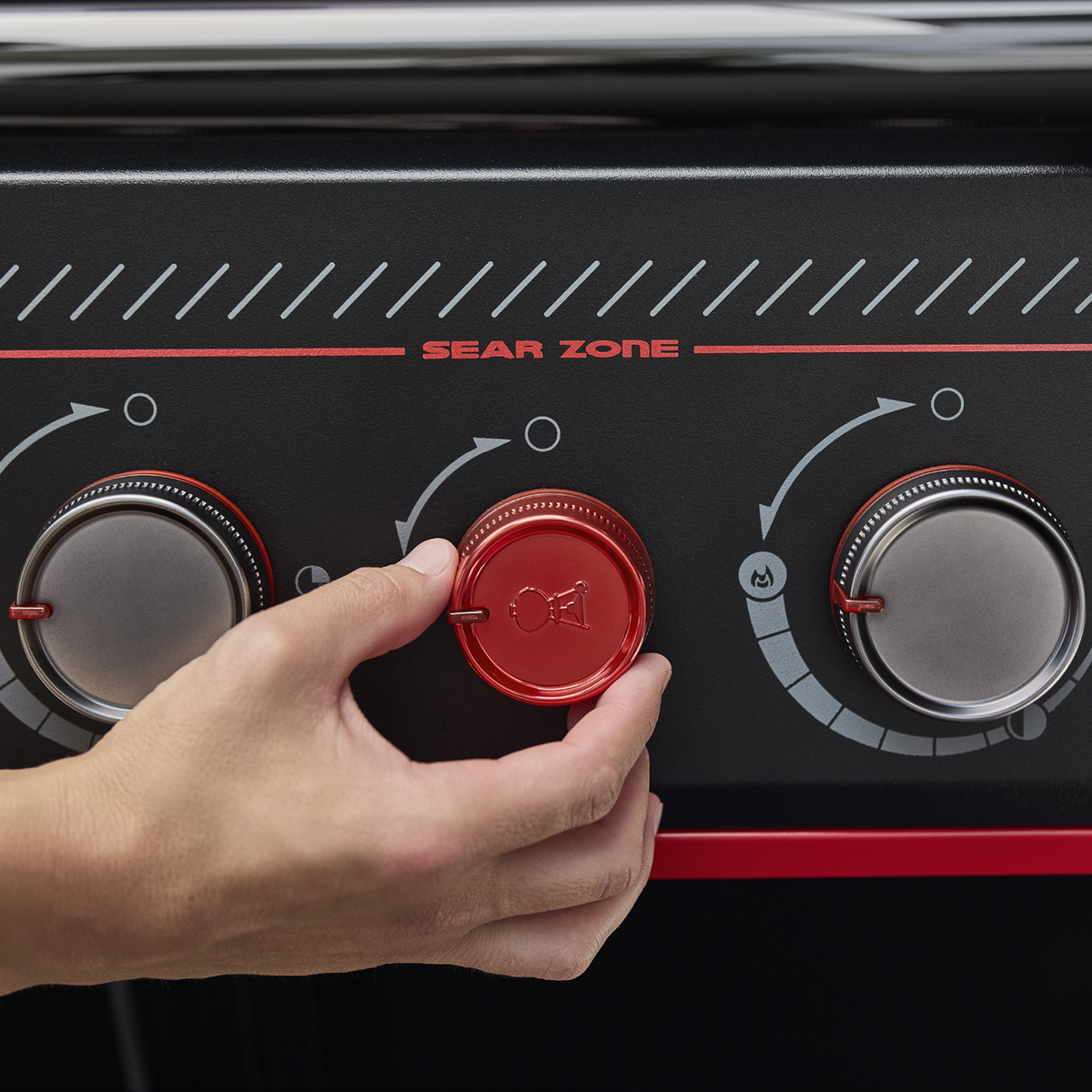 Close-up of a hand adjusting the red Sear Burner knob on the Weber Genesis E-330 Gas Grill Stealth Edition beneath the Sear Zone label.