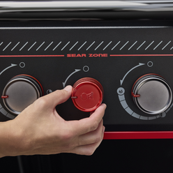 Close-up of a hand adjusting the red Sear Burner knob on the Weber Genesis E-330 Gas Grill Stealth Edition beneath the Sear Zone label.