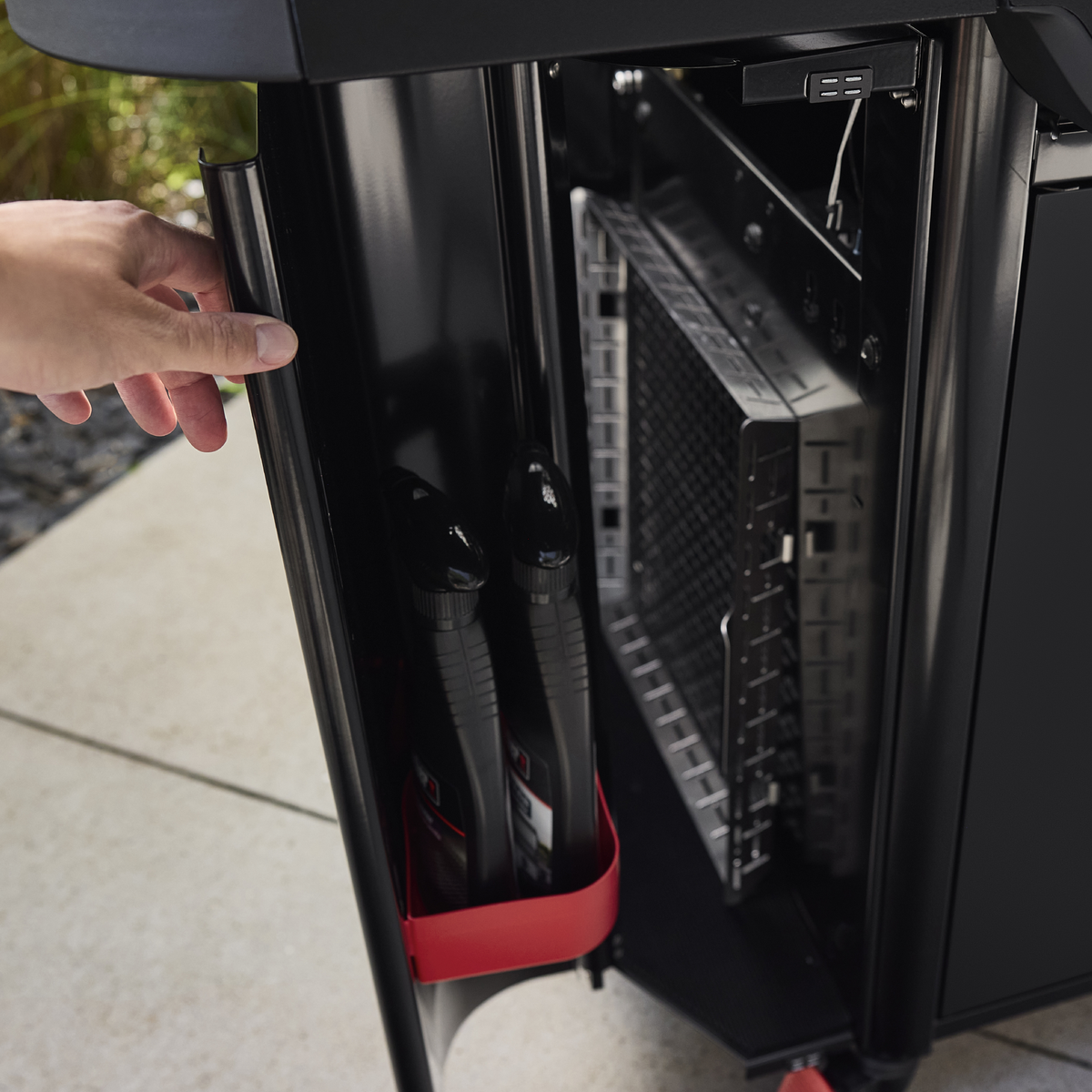 Close-up of a hand opening the grill locker on the Weber Genesis E-330 Gas Grill Stealth Edition, revealing vertically stored cooking accessories and bottles in a red door-mounted holder.