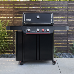 Front view of the matte black Weber Genesis E-330 Gas Grill Stealth Edition on a patio with the lid closed, two side tables, and a red Sear Burner knob, framed by plants and a wooden fence.
