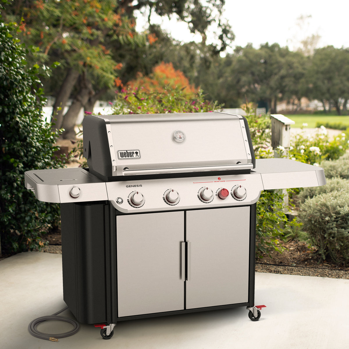 Stainless steel Weber Genesis S-435 gas grill on a garden patio with the lid closed, side shelves, and a double-door storage cabinet.