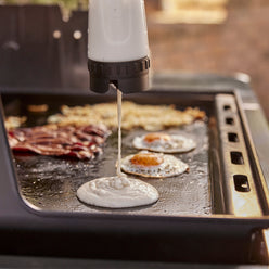 Pancake batter pours onto the Weber Rust-Resistant Griddle Insert while bacon, eggs, and hash browns cook alongside.
