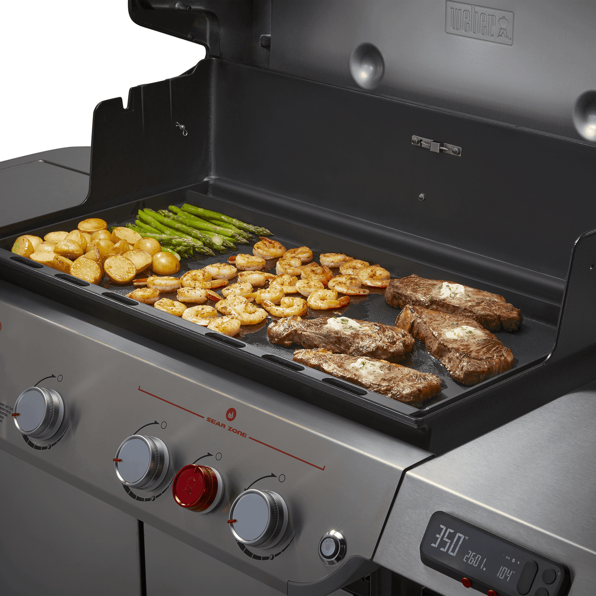 Steaks, shrimp, potatoes, and asparagus cooking together on the Weber Rust-Resistant Griddle Insert inside an open Genesis grill.