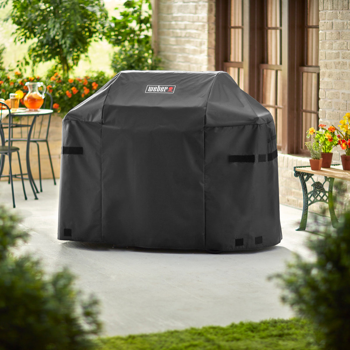 Weber Premium Grill Cover fitted over a Spirit grill on a patio, with side fastening straps and the Weber logo on the front.