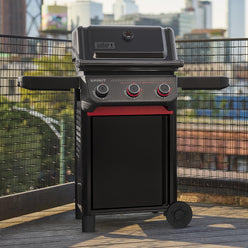 Matte black gas grill on an outdoor deck with a closed lid, built-in thermometer, three control knobs, red accents, and extended side shelves.