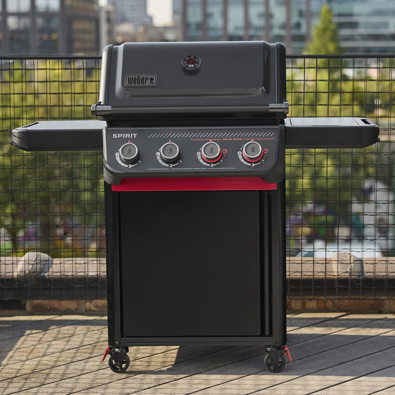 Matte black Weber Spirit E-430 Stealth Edition Gas Grill on an outdoor deck with a closed lid, side shelves, red accents, and city buildings in the background.