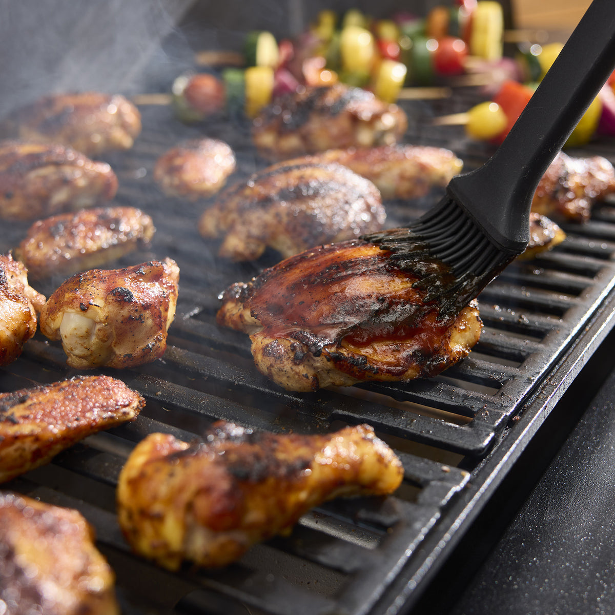 A black basting brush spreads sauce over grilled chicken, with vegetable skewers cooking in the background.