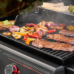 Steaks, bell peppers, and onion wedges cooking on the grill grates, with tortillas on the warming rack and smoke rising from the food.