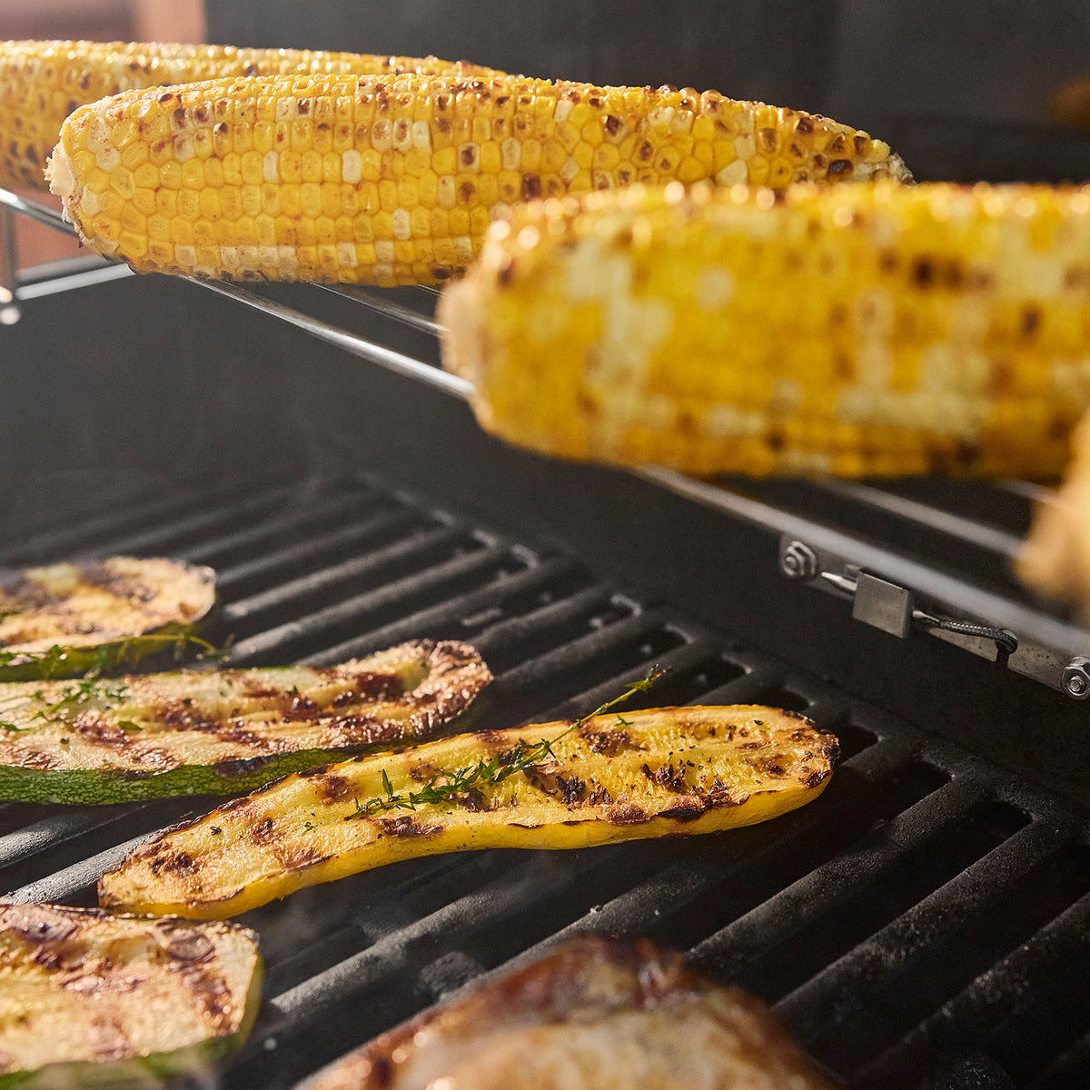 Corn on the cob on the warming rack above zucchini and yellow squash halves with herbs and grill marks on the cooking grates.