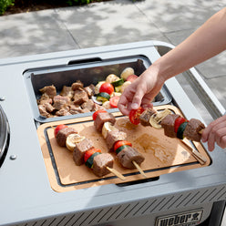 Person assembling beef, tomato, mushroom, and zucchini skewers on a cutting board fitted into the grill’s side table, with a tray of ingredients behind it.