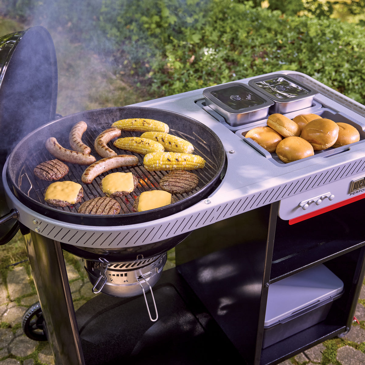 Burgers, sausages, and corn cooking on an open charcoal grill, with buns and lidded food containers in the side table and a storage bin on the lower shelf.