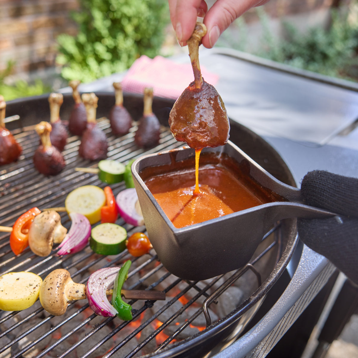 Person lifting a sauce-coated chicken drumstick from a cast-iron saucepan over a charcoal grill, with more drumsticks and vegetable skewers on the cooking grate.