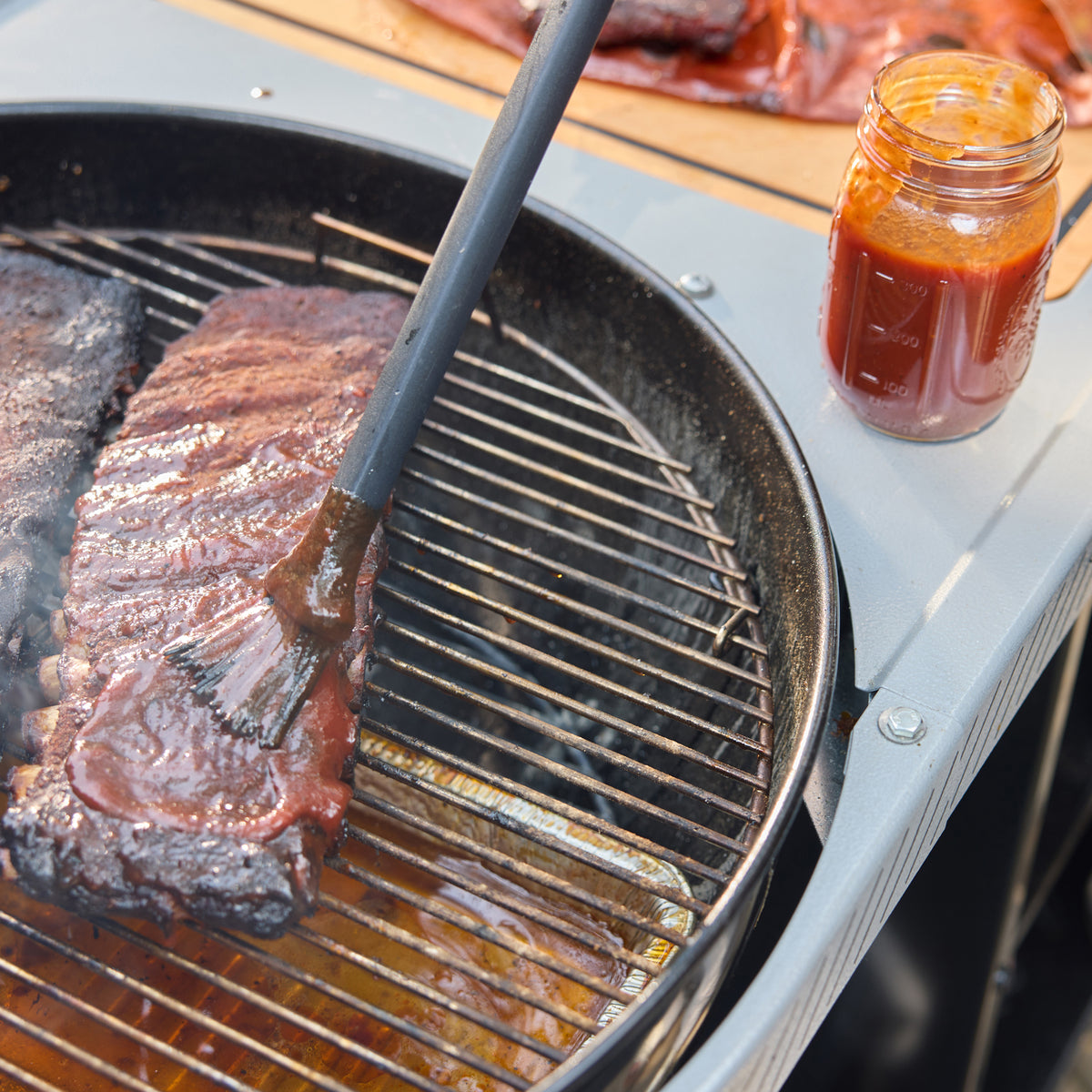 Basting brush spreading barbecue sauce over ribs on a charcoal grill, with a jar of sauce on the side table and a foil drip pan beneath the cooking grate.