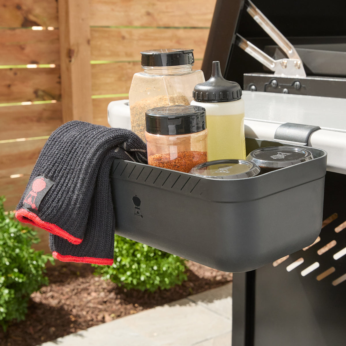 A storage bin attached to the Weber Works side rail on the Weber Slate 36” Rust-Resistant Griddle holds seasonings and a squeeze bottle, with a towel draped over the edge.